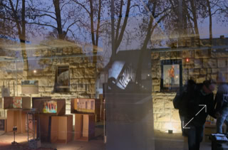 Reflexion eines Schaufensters mit beleuchteten Bildern und zwei Personen, Bäume im Hintergrund.