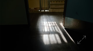 Lichtstrahlen fallen durch ein Fenster und werfen das Schattenornament eines Geländers auf einen dunklen Boden, im Hintergrund ist eine Treppe sichtbar.