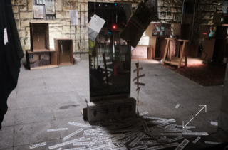 Ein Raum mit einem Spiegel in der Mitte, umgeben von verstreuten Papierstreifen auf dem Boden; im Hintergrund sind Kartons an einer Steinwand zu sehen.