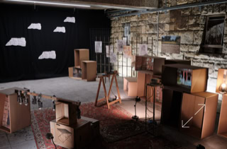 Ein Raum mit Kartons, Teppichboden, einer schwarzen Wand mit weißen Schriftstücken und einer Steinwand im Hintergrund.