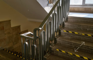 Ein Treppenhaus mit Metallgeländer und gelb-schwarzen Markierungen auf den Stufen, beleuchtet durch ein Fenster im Hintergrund.