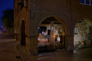 Ein beleuchteter Raum mit offenen Bögen, gefüllt mit gestapelten Kartons, daneben ein dunkler Gehweg.
