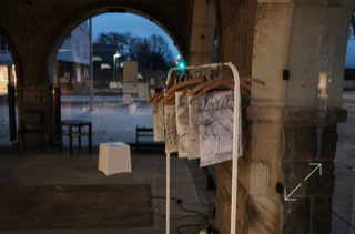 Kleiderständer mit aufgehängten Zeichnungen unter einem Bogen in einem überdachten Bereich, im Hintergrund eine Straße und Bäume.